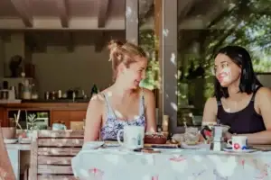 Δύο νέες γυναίκες απολαμβάνουν καφέ σε εξοχική κουζίνα, χαμογελαστές και υγιείς χωρίς συμπτώματα λοίμωξης χάρη στην ενίσχυση του ανοσοποιητικού με ομοιοπαθητική στην Αθήνα