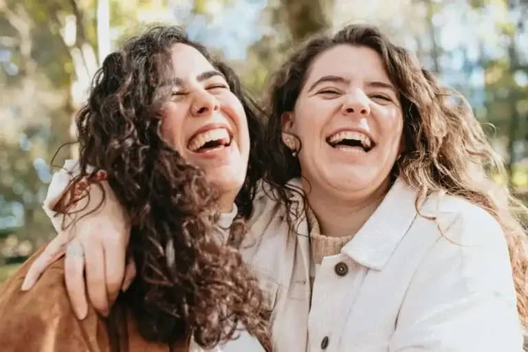 Δύο νέες γυναίκες γελούν αγκαλιασμένες με γνήσια χαρά χωρίς ίχνη ευερεθιστότητας από προεμμηνορρυσιακό σύνδρομο χάρη στην ομοιοπαθητική θεραπεία στην Αθήνα