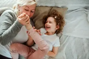 Γιαγιά παίζει χαρούμενα με την εγγονή της στο κρεβάτι, κρατώντας τα χέρια της χωρίς πόνο ή δυσκαμψία από ρευματοειδή αρθρίτιδα χάρη στην ομοιοπαθητική θεραπεία στην Αθήνα