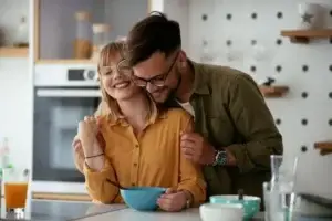 Ζευγάρι 30-35 ετών σε στιγμή τρυφερότητας στην κουζίνα του σπιτιού τους, η γυναίκα με μουσταρδί πουκάμισο χαμογελά καθώς ο σύντροφός της την αγκαλιάζει, συμβολίζοντας την ελπίδα και τη σύνδεση που βοηθούν στη φυσική γονιμότητα