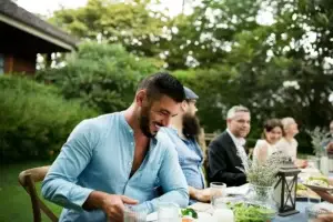 Χαρούμενος άνδρας απολαμβάνει το φαγητό του με παρέα σε εξωτερικό χώρο, συμβολίζοντας την ανακούφιση από τον πόνο του πεπτικού έλκους και την καλή πέψη μέσω της ομοιοπαθητικής.