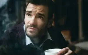 Άνδρας κοιτάζει ανήσυχος έξω από παράθυρο κρατώντας κούπα, απεικονίζοντας την εσωτερική ανασφάλεια του τύπου Lycopodium.